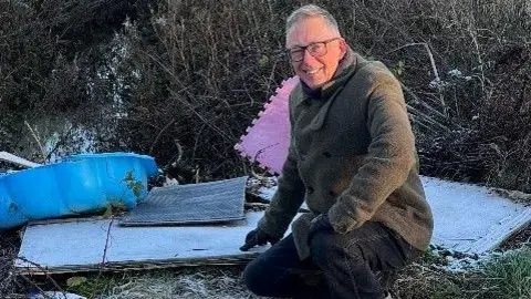 Phillip Redshaw Phillip is wearing a light green jacket and black gloves. He has grey hair and dark-rimmed glasses and is bending down, smiling at the camera at the site of fly-tipped rubbish. Included in the rubbish is a plastic blue children's bathing pool and pink mat