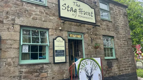 The exterior of the pub. It is a granite building with green painted window frames and door frame. There is a sign above the entrance which reads The Stag Hunt at Ponsanooth. Above the doorway is a sign which reads ENTRANCE and to the left a branded sign reads GOOD FOOD, BED AND BREAKFAST and A WARM WELCOME. In front of the entrance is a sign which reads SAVE THE STAG.