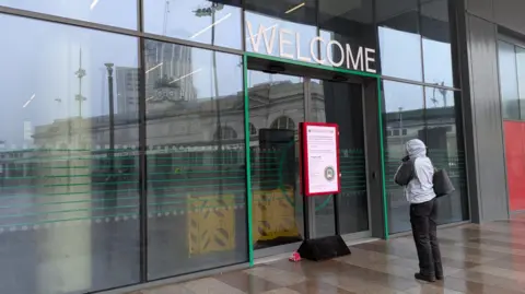 BBC A person wearing a raincoat looks at a sign in front of the Cardiff Bus Interchange. The sign has a red frame. Behind it are closed glass doors and a sign that says Welcome.