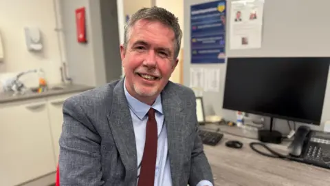 Headteacher Richard Blackburn smiling in a suit in his office. He is wearing a burgundy tie