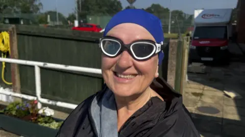 A woman with a black top, black goggles and a blue cap standing outdoors in front of a wooden fence