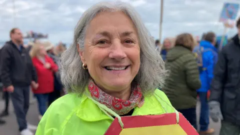 A woman with white hair, wearing a pink and white patterned gaitor around her neck and a high-visibility jacket. She's smiling at the camera and there's a crowd of people behind her.