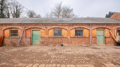 Delapre Abbey Stable buildings, with windows in them, two green doors, an area with stones on the ground and wood to the right. 