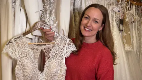 Bronwen Barclay is smiling and holding her wedding gown. Bronwen has long brown hair and is wearing a red crew neck jumper. The top of the dress on a hanger is visible. It is short sleeved, ivory coloured and made of lace. There are a variety of wedding dresses hanging behind her. 