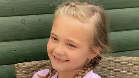 A blonde girl is smiling. Her hair is in plaits and she is wearing a pink top. She sits on a wicker chair in front of a wooden structure painted green.