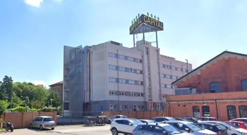 A wide exterior image of The Hotel Capital. It is a large grey building with a large sign on top of the roof which says Capital in yellow letters. Above the sign are four yellow stars. 