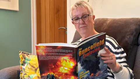 Eric Johnson/BBC A woman wearing a black and white stripy top reads a book about Buncefield, with vivid images of the explosion on the front and back covers.