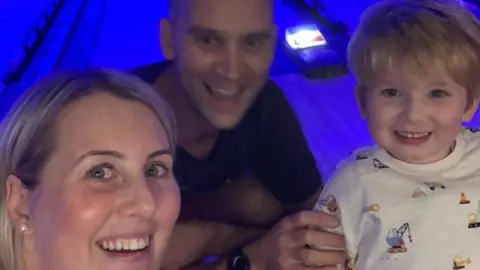 Family handout A close-up selfie of a blonde woman and a man next to a young boy with blonde hair. The boy is smiling and wearing a top with cranes on it. They appear to be at a bowling alley and the lighting is blue.