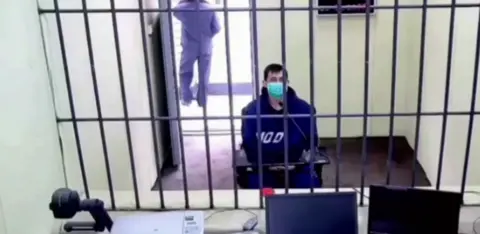 A man wearing a hoodie and a surgical mask sits behind a metal mesh in a room. There are two computer monitors on a desk in front of the mesh. Behind him, the door is open and a person is standing outside the door.