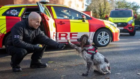 WMFS A brown and white dappled cocker spaniel sits on the ground and holds her paw up towards a man kneeling down. Two fire service vehicles are behind them.