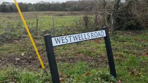 A road sign partially leaning with the words "Westwells Road" written on it. Behind it is a fence and a field and a tree line in the far background.