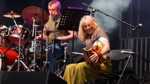 Belfast City Council A woman on a stage is sitting on a chair and holding an accordion. There is a drummer in the background. She is smiling. 