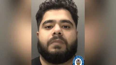 West Midlands Police A mug shot of a man with a moustache and dark, bushy beard. He has short curly hair and brown eyes. The top of his black t-shirt can be seen.