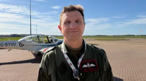 Pilot, wearing a khaki green flight suit, is stood in front of a white aircraft. Blue sky with patchy clouds in the background.