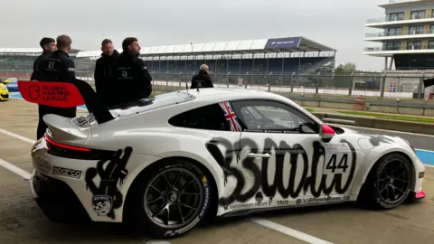 A white Porsche touring car stands in the paddock at Silverstone, with team members in black race suits standing nearby and grandstands in the background. The car is white with red spoilers and a Union flag logo. On the side is sprayed $33,000,000.