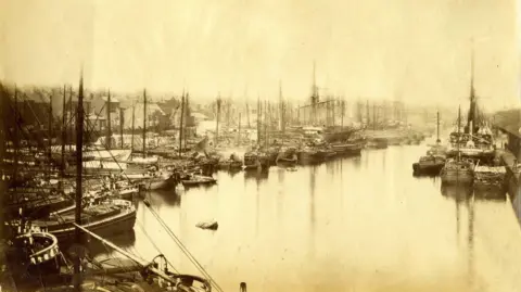 Hull History Centre A sepia-tinged black and white photo of a dock, perhaps taken in the early years of the 20th Century, showing dozens of boats with masts tightly packed to the left. To the right, a larger ship is moored against a quay.