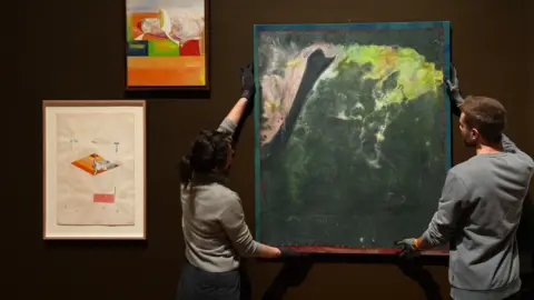 PA Media A large painting is being held up by two people on the right. It mainly includes different shades of green. There are two smaller pieces of art work hanging on the wall on the left.