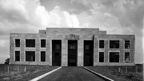 A black and white photo depicts a two story building made from stone. It has three columns in the middle and a crest over the front entrance.