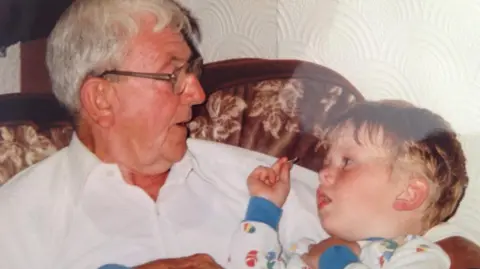 Family photograph The late Robert Worthington with white hair and wearing glasses and a white shirt holds his grandson Adam Salisbury, then a toddler, with blond hair who is sitting on his knee wearing blue and white pyjamas on. 