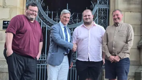 Group photo of SDP party members alongside Rhys Carr. Rhys is wearing a pink short-sleeved shirt and is shaking hands with a fellow member of the SDP. They are smiling.