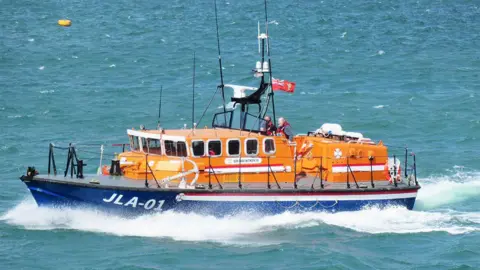 Jersey Lifeboat Association The Sir Max Aitken III underway on a sunny day in calm waters. The sea looks turquoise and the boat is creating a white bow wave. There are two men at the helm, both wearing lifejackets.