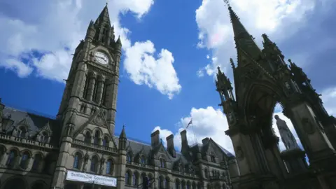 A photograph of Manchester Town Hall, which was taken in 2020, and shows the front of the building including its clock and a white banner which says 'sustainable government' and a year which is not identifiable. In the background is a blue sky with some clouds.