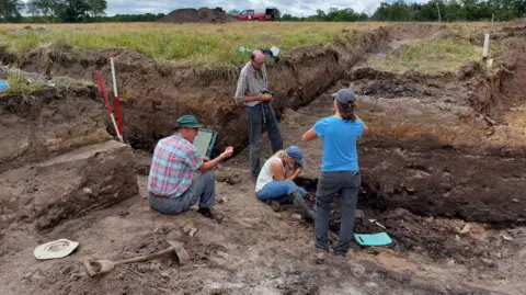 Carl Sayers/NWT Men and women taking part in a dig in a field to excavate an ancient Ice Age pond. They stand in deep trenches at least five feet high (150cms). A truck and a mound of soil can be seen in the distance.