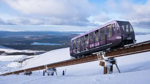 The funicular railway is a long metal rail installed on a mountainside. A purple, railway carriage-like vehicle travels along the rail. The ground below is covered in snow and beyond is sweeping landscape of hills and lochs.