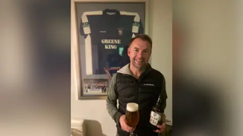 Contributed Marcus Stewart is pictured smiling at the camera while holding a beer bottle in one hand and a glass filled with the beer in another. He is wearing a black gilet jacket with a green hoodie underneath. An old Ipswich Town football shirt in blue and white is framed on the wall behind him.