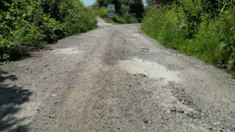 A close up image of a gravel pot hole ridden road. It has patches of dirt and small rocks, set in a rural lane. There is also a sign to the football club in the distance.