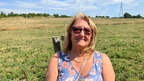 Photograph of Joan Higson from Atherton. She is pictured in front of farmland, with an electricity pylon and trees in the background. 