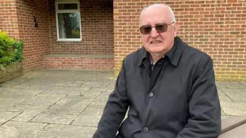 A man is sitting on a bench outside a village hall. He is wearing a black coat and sunglasses