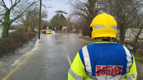 A fire safety officer in hi-viz and a yellow helmet watches firefighters in waders rescue occupants of a white car stuck on a flooded road