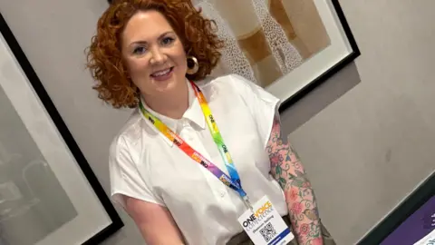 Equity Shannon from Equity is smiling. She has ginger hair and a sleeve tattoo on her left arm. Her tattoos are all flowers. She has a white shirt and a multicoloured lanyard.