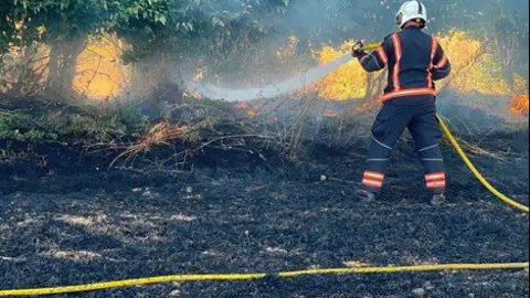 A fire fighter in a black uniform, putting out a field fire with a yellow hosepipe.