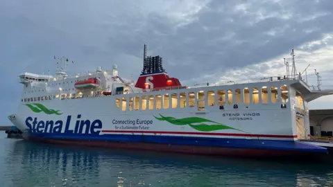 The Stena Vinga ferry docked at the St Helier Harbour. It is a large, white vessel with the words 'Stena Line' written in blue on its side. In smaller letters, it says 'Connecting Europe for a Sustainable Future'. There is also a light green logo and a red line which runs towards the back of the vessel.