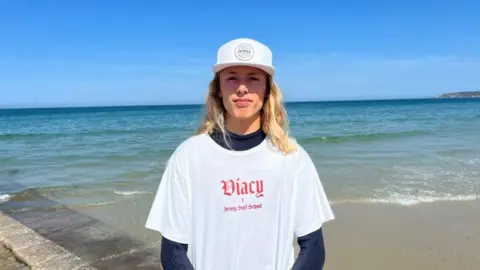 Jesse Heddercott is 16-year-old boy with long, blond hair past his shoulders. He is standing on a slipway. He wears a white cap, and a white T-shirt over a wetsuit that says Jersey Surf School. The sea is covering some of the slip, and is turquoise in the shallows before turning darker further out. The sky is blue.