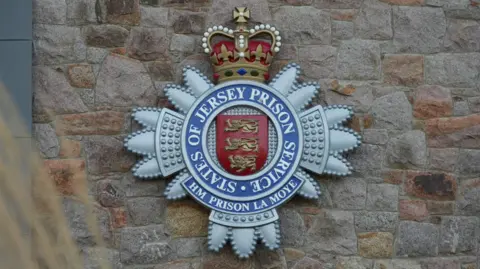 A picture of a logo that is on the outside of the prison building. It is a silver, red and blue logo that reads 'HM PRISON LA MOYE'.