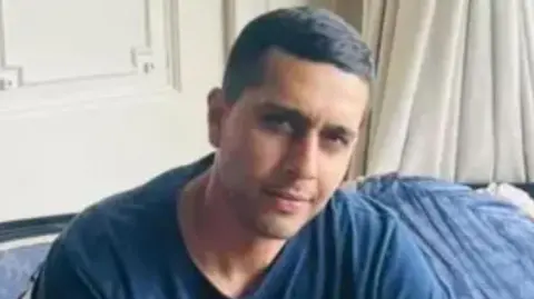 Kamran Aman pictured sitting on a blue sofa in living room with white curtains behind and white wood panelling on the wall. He has short dark hair and wears a  blue t-shirt