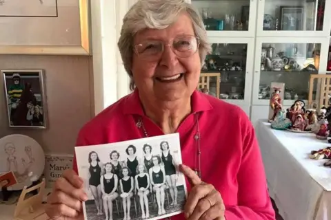 Scottish Swimming Mary Black when she was in her 80s. She has short grey hair and glasses and is wearing a pink jumper. She is holding an old photograph of her synchronised swimming team from when she was a teenager. There are figurines and ornaments in the background.
