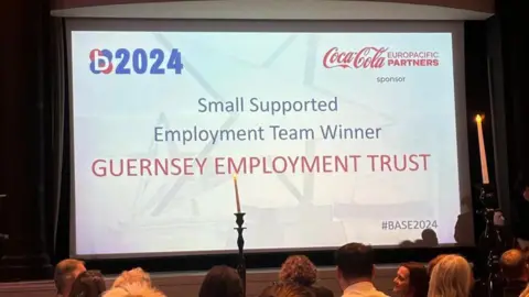 Elaine Strappini The screen at the awards ceremony which has '2024' in the top left corner. At the centre are the words 'Small Supported Employment Team Winner Guernsey Employment Trust'. In the bottom right corners are the words '#BASE2024'.