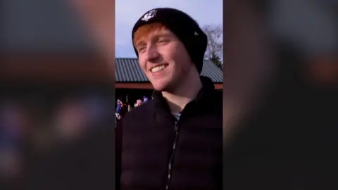 A man, Angry Ginge, smiling outside wearing a beanie hat and black jacket.