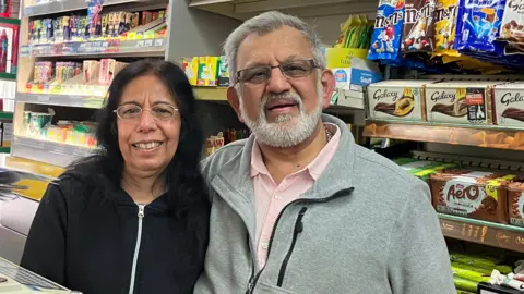 Smitty is on the left of picture. She is wearing a vary dark blue zip-up top and has black, shoulder length hair. Charlie is standing next to her. He is wearing a grey top over a pink shirt. He has a grey beard and hair. Both are wearing glasses and standing behind the counter of their shop.