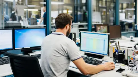 A man with a grey polo shirt, dark hair and dark brown facial hair sits at a desk with his back to the camera. He is sitting on a black chair and there are three computers in front of him, he is working on one. He appears to be in an office setting with other colleagues working in the background.
