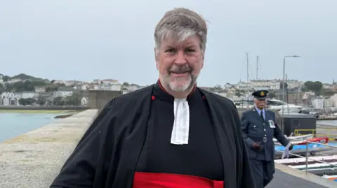 Rev Tim Barker standing next to a wall. He is wearing a black outfit with red accents and a white clerical collar. In the background there is a man wearing a blue suit with military medals