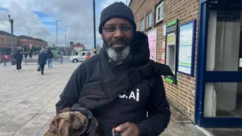 Julita Waleskiewicz/BBC Howard pictured outside Luton train station on the bus station concourse, with lamp posts and people behind him. His is holding a brown speckled dog and wears a black beanie hat, glasses, a black hooded jacket and the black strap of a bag across his chest.