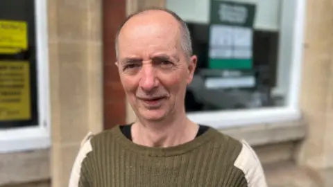 A man with grey hair and a green top standing in front of a building