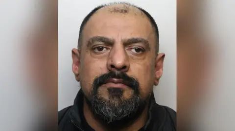 Police custody image of Suaiel Haider. He has receding black hair and a beard and is wearing a black shirt.