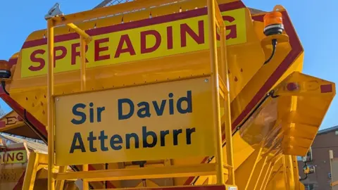 Yellow gritting vehicle with Sir David Attenbrrr on its rear.