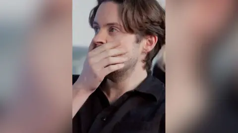 A man with short, brown hair covers his mouth with his hand